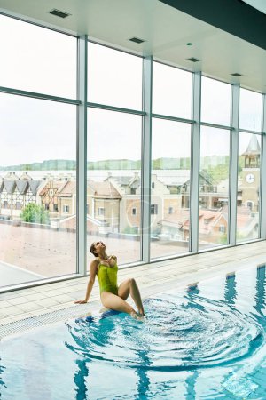 Photo for A young brunette woman in a green bathing suit sits gracefully on the edge of an indoor swimming pool. - Royalty Free Image