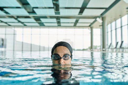 Young woman glides through pool in goggles and swim cap, embodying fluid motion and tranquility.