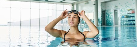 Téléchargez les photos : Une jeune femme en maillot de bain et bonnet de bain glisse gracieusement dans l'eau, portant des lunettes dans un spa intérieur serein. - en image libre de droit