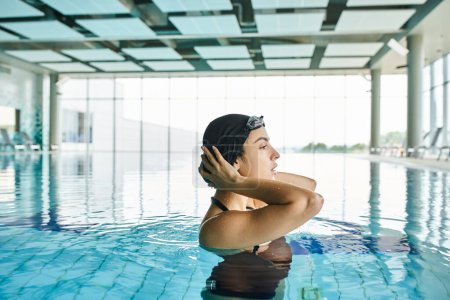Eine junge Frau in Badeanzug und Badekappe schwimmt anmutig in einem Indoor-Spa, trägt eine Brille.
