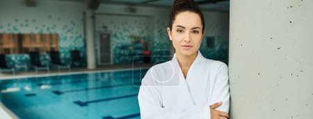 Téléchargez les photos : Une jeune, belle femme brune debout élégamment à côté d'une piscine intérieure dans un cadre spa. - en image libre de droit