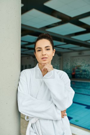 A young woman in a bathrobe enjoys a peaceful moment by an indoor pool in a spa setting.