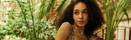 young african american woman with curly hair in animal print look in a tropical setting, banner
