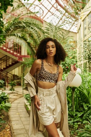 young african american woman with curly hair standing in trendy look and boots in tropical setting