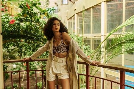 Chic african american woman with curly hair posing in her stylish look in tropical conservatory