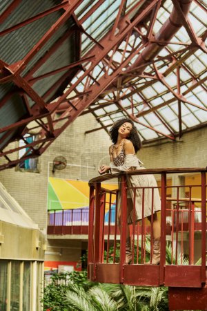 curly and chic african american woman in animal print look posing against industrial backdrop