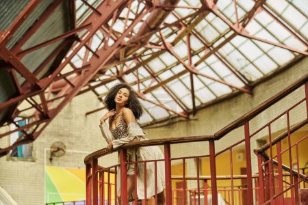 african american woman with curly hair in animal print look posing against industrial backdrop