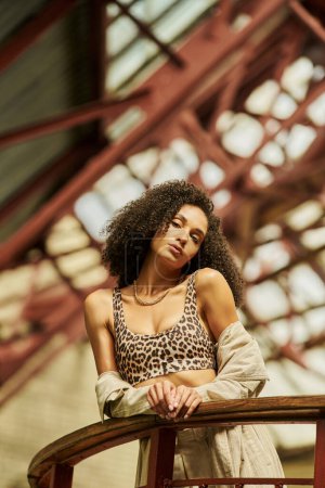 curly young african american woman in animal print look posing against industrial backdrop