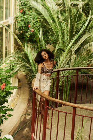 Modische junge schwarze Frau mit lockigem Haar posiert auf einer Brücke im Garten