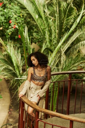 Téléchargez les photos : Jeune femme noire à la mode avec des cheveux bouclés posant sur un pont métallique dans un cadre de jardin luxuriant - en image libre de droit