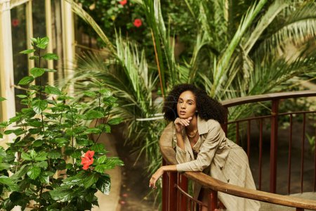 stylish young black woman with curly hair leaning on metallic structure in green garden setting