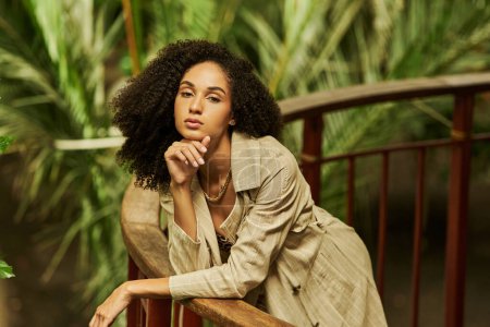 african american woman with curly hair leaning on metallic structure in green garden setting