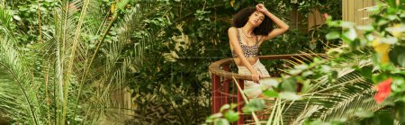 Fashionable african american woman with curly hair posing in a lush tropical environment, banner