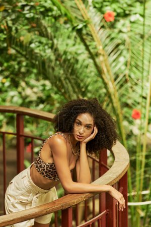 hübsche afrikanisch-amerikanische Frau posiert in trendiger Kleidung auf metallischer Brücke in grünem botanischem Ambiente