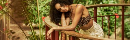 flirty african american woman with curly hair posing on metallic bridge in green setting, banner