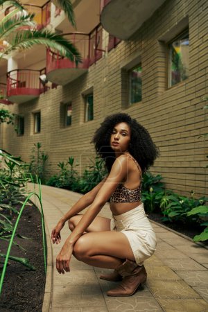 Confident black woman in chic style attire and boots posing in atrium with botanical backdrop