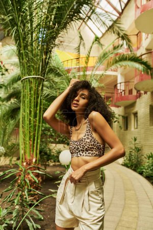 black woman in stylish animal print attire posing with hand near curly hair in atrium with plants