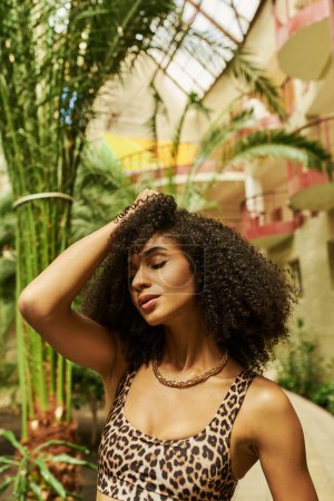 black woman in animal print attire posing with hand near curly hair in atrium with green plants