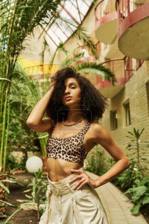 black woman in animal print attire posing with hand near curly hair in botanical atrium with plants