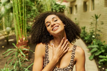 happy black woman in animal print top posing with hand near neck in atrium with green plants
