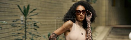 curly young black woman in dress and sunglasses poses in animal print gloves in urban garden, banner
