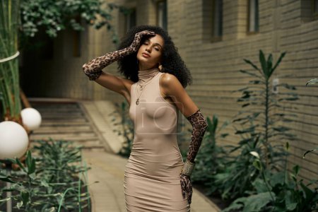 Sophisticated and curly black woman in dress and animal print gloves posing in urban garden