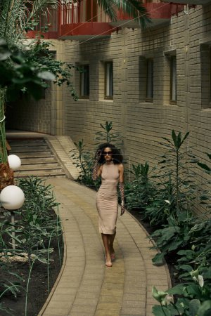 elegant african american woman in dress animal print gloves and sunglasses walking in urban garden
