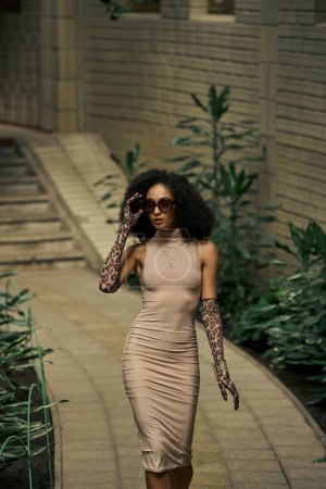 Foto de Elegante mujer negra en vestido, guantes de impresión animal y gafas de sol caminando en el jardín urbano - Imagen libre de derechos