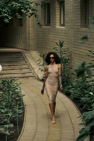 curly black woman in elegant dress, animal print gloves and sunglasses walking in urban garden