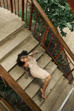 curly black woman in dress and animal print gloves lying on stairs in urban garden, top view