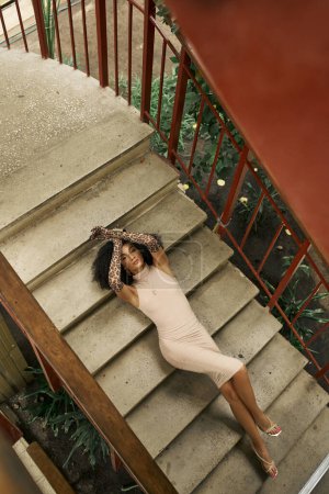 jeune femme noire en robe et gants imprimés animaux couchés sur les escaliers dans le jardin urbain, vue sur le dessus
