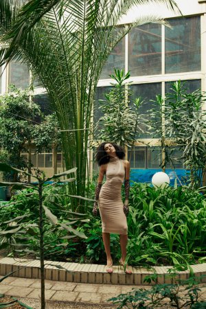 longitud completa de la mujer afroamericana en vestido y guantes de impresión animal posando entre las plantas en el jardín