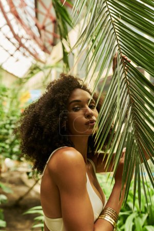 charming african american woman in swimsuit posing with green palm leaf in a lush greenhouse