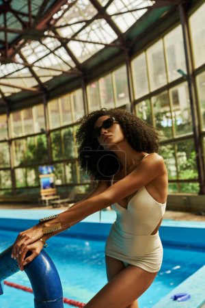 Téléchargez les photos : Élégante femme noire en maillot de bain chic et lunettes de soleil tendance debout au bord de la piscine, mode vacances - en image libre de droit