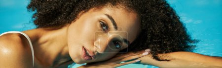 portrait of african american woman with curly hair looking at camera and swimming in water, banner