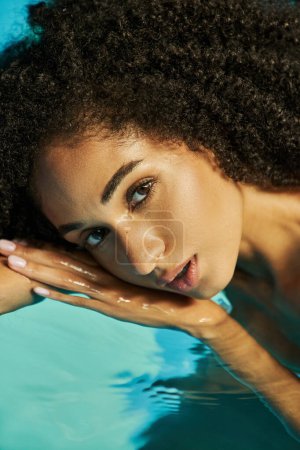 Porträt einer hübschen afrikanisch-amerikanischen Frau mit lockigem Haar, die in die Kamera schaut und im Wasser schwimmt