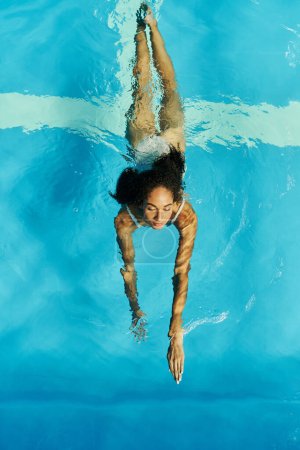 Téléchargez les photos : Vue de dessus de la femme afro-américaine frisée nageant dans l'eau claire et bleue de la piscine, sereine et paisible - en image libre de droit
