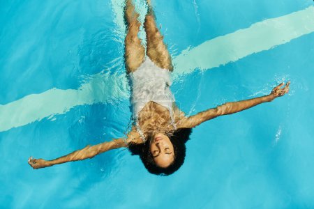 Téléchargez les photos : Vue du dessus de la jeune femme afro-américaine nageant sur le dos dans l'eau claire de la piscine, sereine et paisible - en image libre de droit