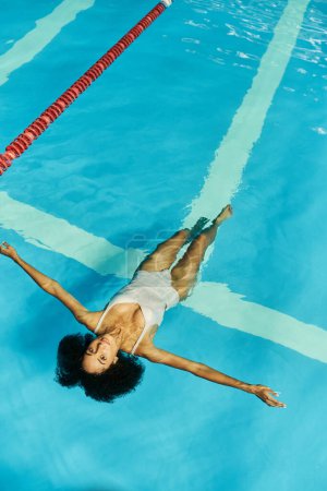 Téléchargez les photos : Vue du dessus de la jeune femme afro-américaine nageant sur le dos dans l'eau bleue de la piscine, tranquillité - en image libre de droit
