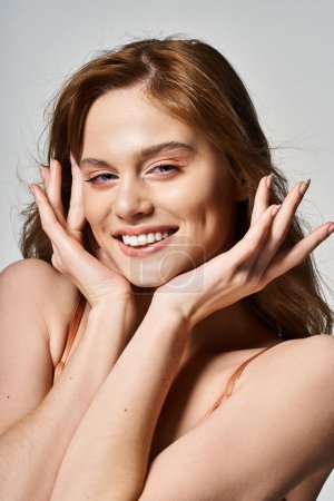 Cheerful smiling woman with trendy makeup, looking at camera, hands near face on grey background