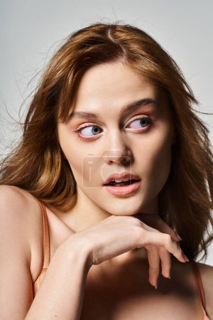 Young surprised woman with trendy peachy makeup, looking away, hands near face on grey background