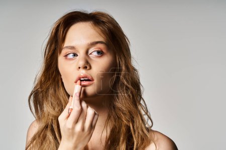 Téléchargez les photos : Portrait de beauté de femme détournant les yeux avec un maquillage nu appliquant du rouge à lèvres sur fond gris - en image libre de droit