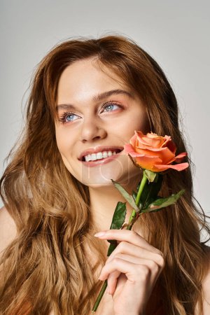 Foto de Hermosa mujer joven sonriente con ojos azules, con rosa melocotón cerca de la cara sobre fondo gris - Imagen libre de derechos