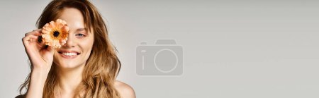 Beautiful smiling young woman with gerbera flower near eye on grey background, banner