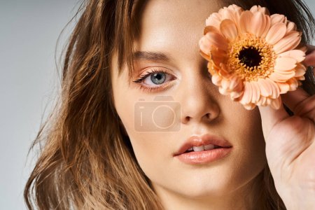 Closeup beauty shot of pretty woman with peach makeup and gerbera daisy near eye on grey background
