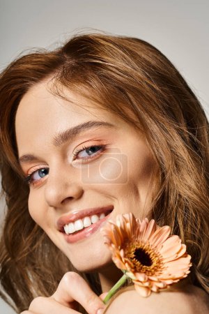 Closeup beauty shot of smiling woman with peach makeup and gerbera daisy on grey background