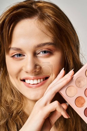 Portrait of a beautiful smiling woman holding nude eyeshadow palette near face and looking at camera