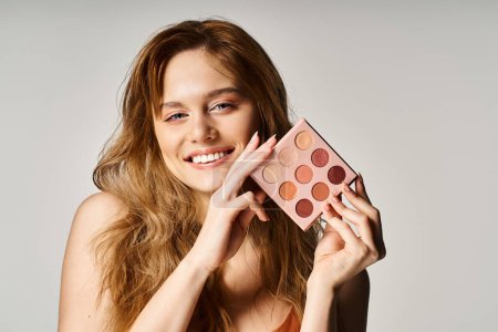 Beauty shot of smiling woman holding nude eyeshadow palette near face and looking at camera