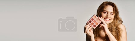 Téléchargez les photos : Photo de beauté d'une femme souriante tenant une palette de fards à paupières nus près du visage et regardant la caméra, la bannière - en image libre de droit
