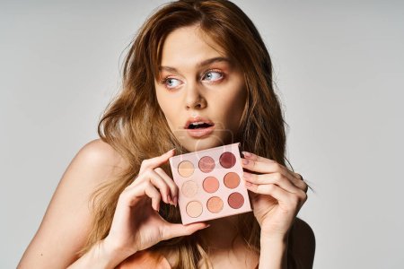 Beauty photo of surprised woman holding nude eyeshadow palette near face and looking away